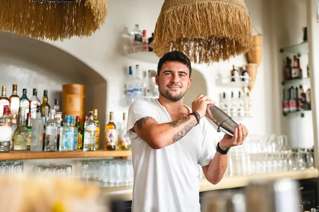 Tattooed bartender shaking cocktail shaker behind bar in upscale restaurant with liquor bottles on shelf.