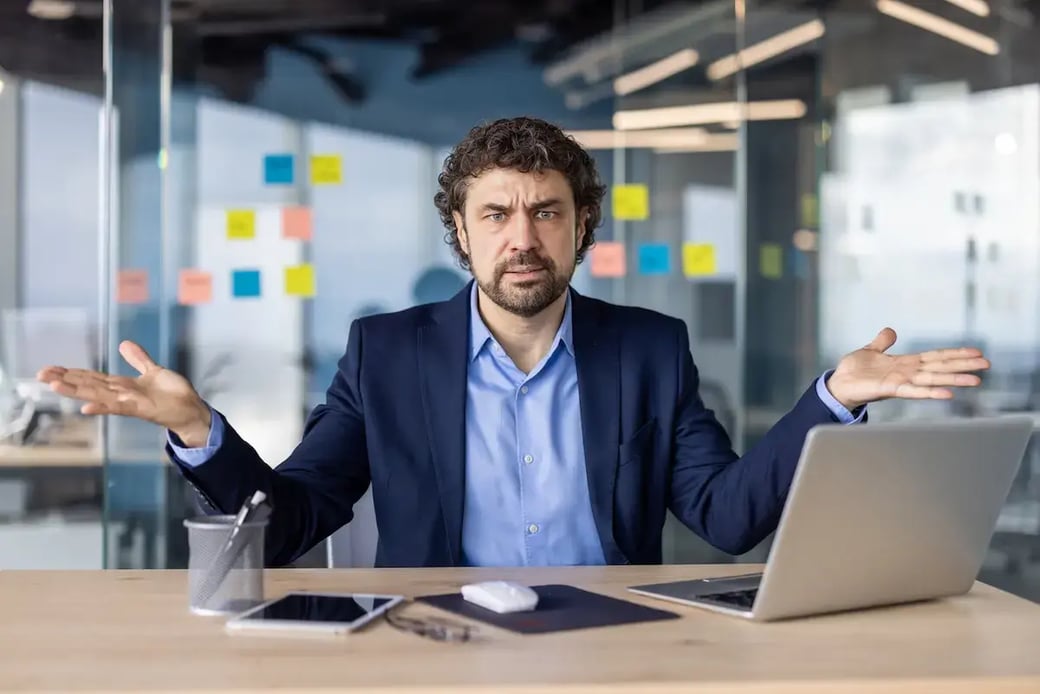 Confused businessman sitting at desk with hands raised, surrounded by office tech and sticky notes on a glass wall.