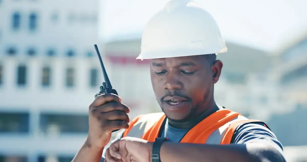 Construction supervisor in hard hat using walkie-talkie and checking watch on job site.