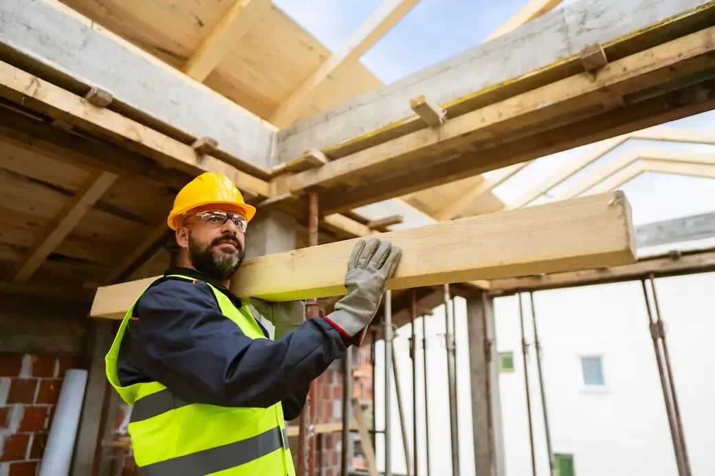 Construction worker in hard hat and safety vest carrying wood beam at active job site under roof framing.