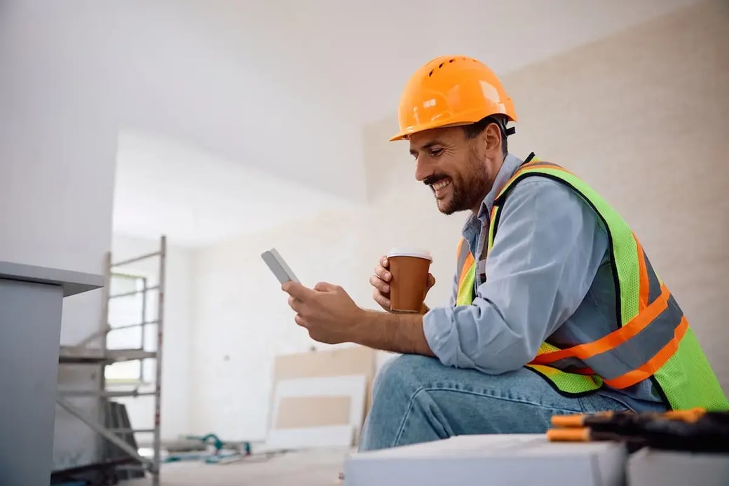 Smiling construction worker in safety vest and hard hat checking phone while holding coffee.