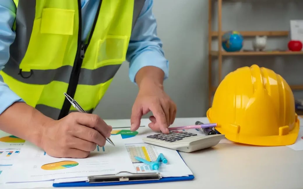 Construction worker in safety vest reviewing project finances with charts, calculator, and hard hat on desk.