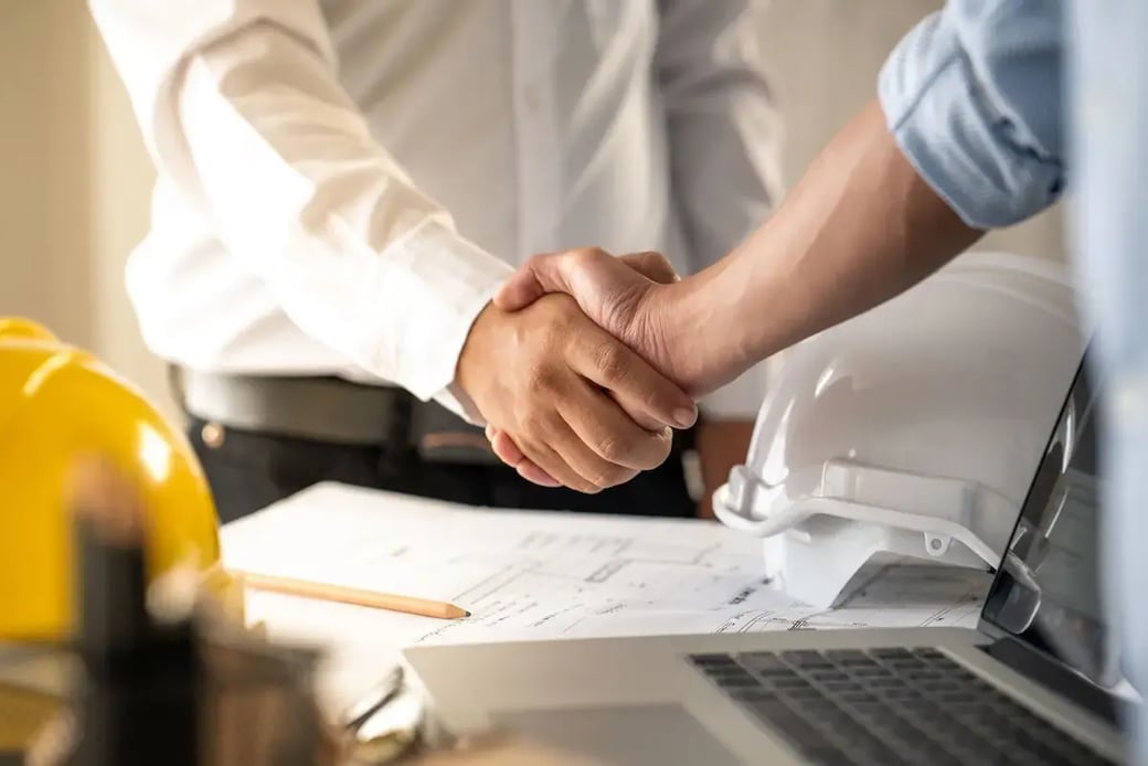 Construction workers shaking hands at job site over blueprints, symbolizing trust and financial planning success in contractor partnerships.