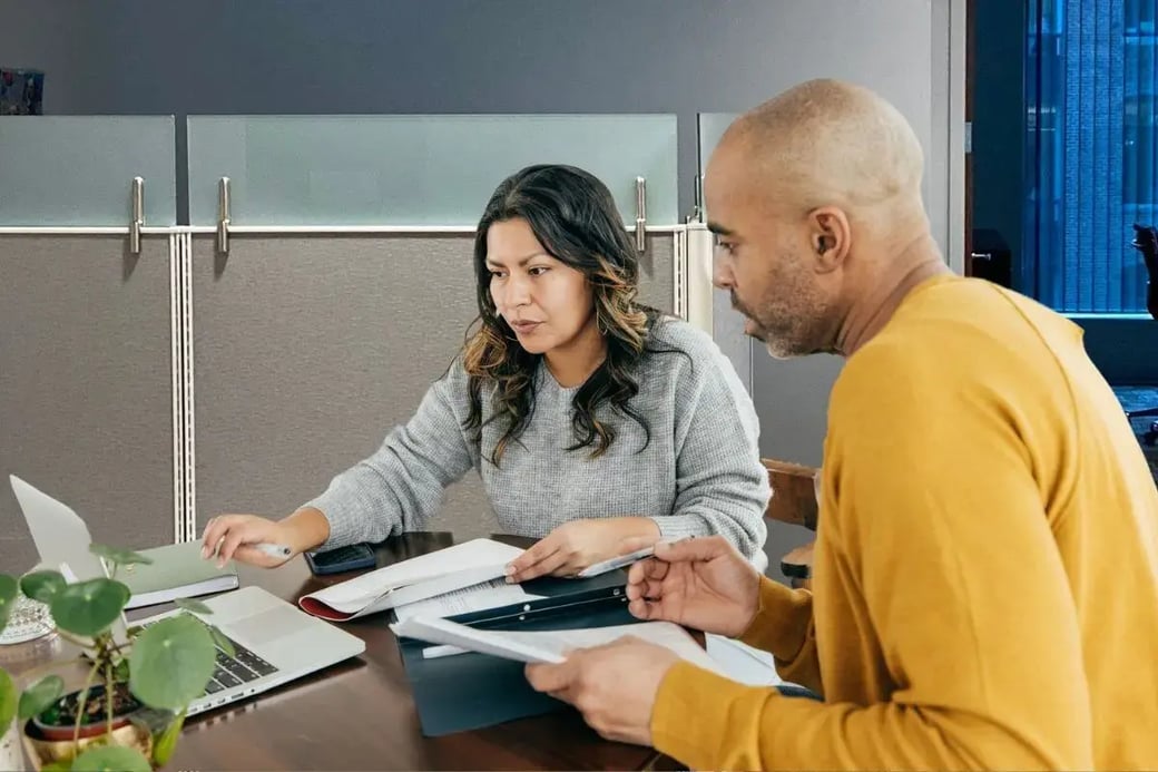 Businesswoman and businessman reviewing financial documents and working on a laptop at a shared office desk with privacy dividers and natural light.