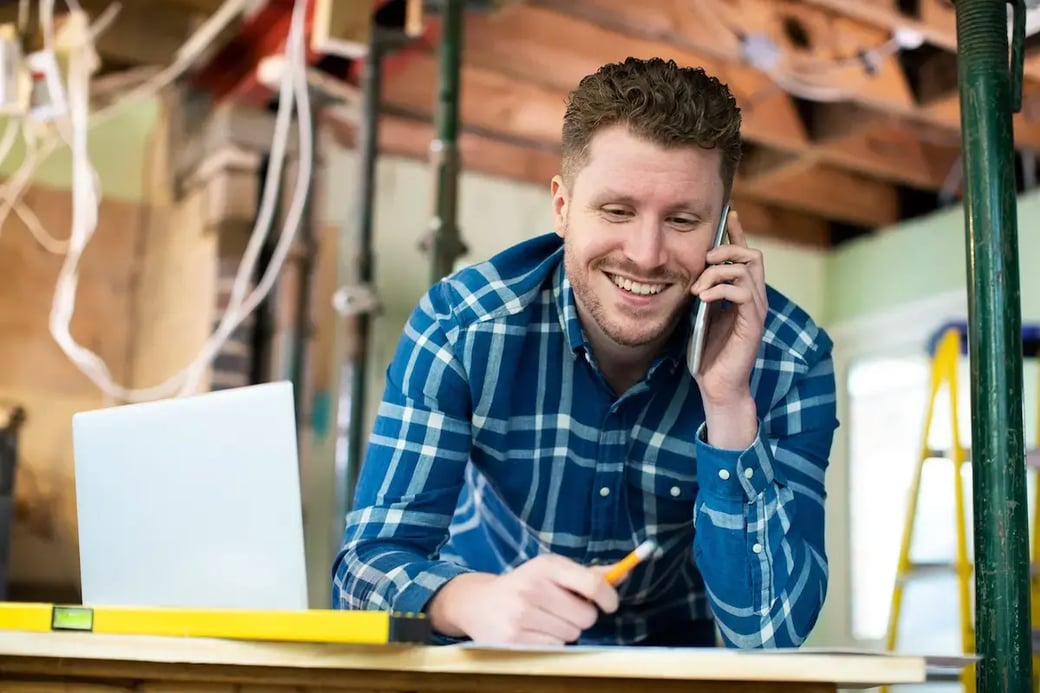 Smiling contractor speaking on smartphone while reviewing renovation plans at indoor construction site.