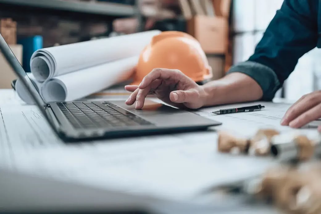 A contractor working on laptop with blueprints and hard hat visible.