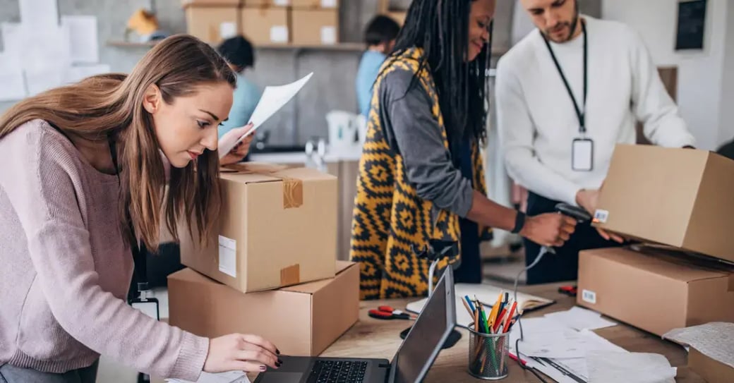 Three warehouse workers process and prepare customer orders, scanning and labeling cardboard boxes for shipment on a desk with a laptop and office supplies.