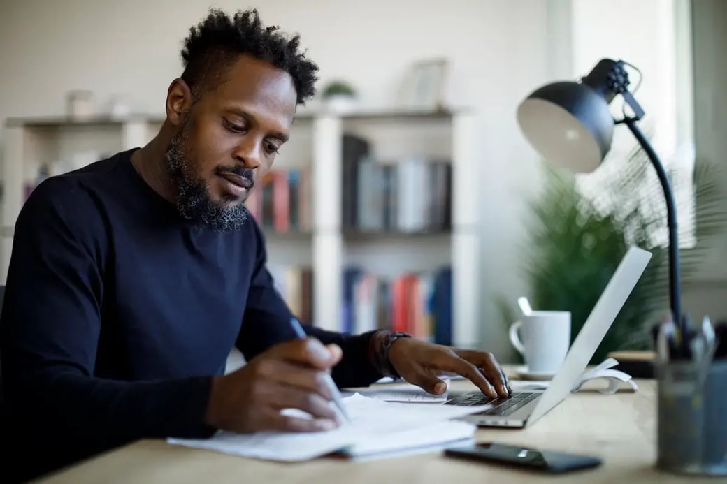 Man working remotely at a desk with a laptop, reviewing paperwork for business or license application.