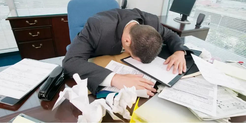 Overwhelmed man in business attire with head on desk, surrounded by disorganized paperwork and office clutter.