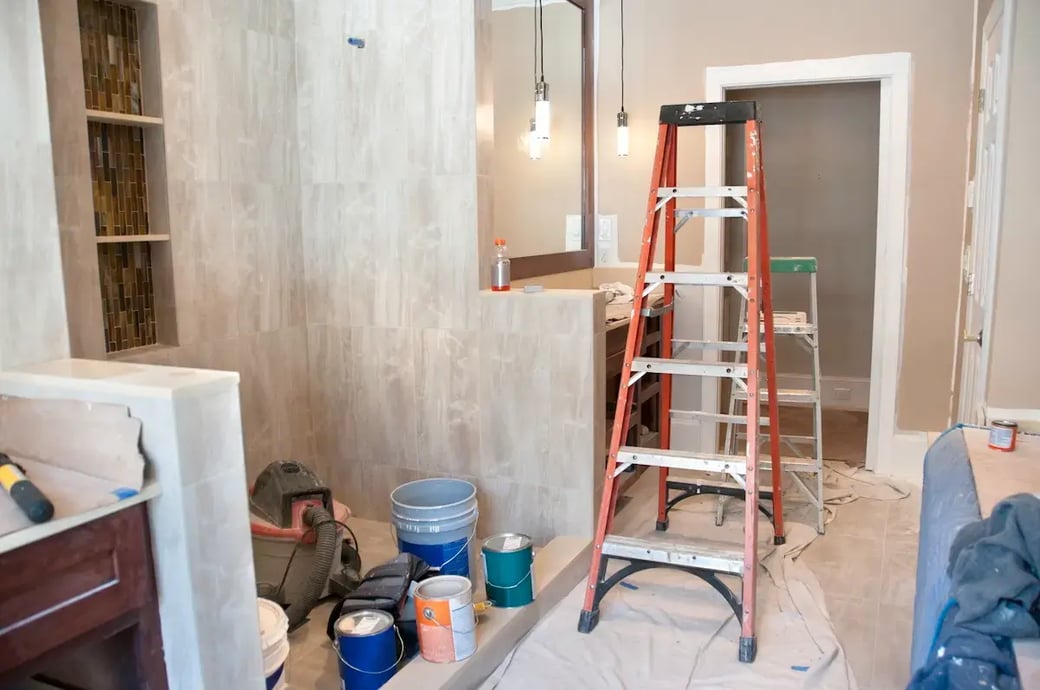 Bathroom renovation in progress with buckets, tools, and ladder on plastic-covered floor in residential home remodeling project.