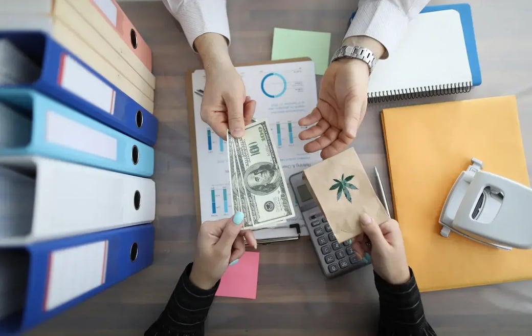 Close-up of financial exchange with cash for cannabis in an office setting with charts and binders.