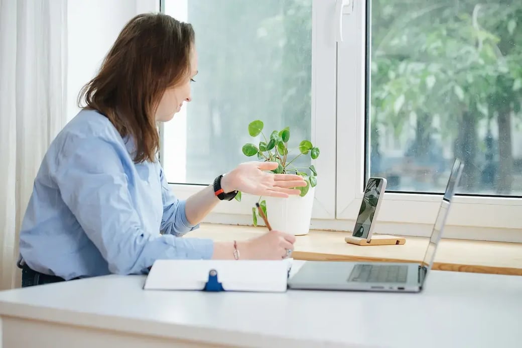 Woman having video call on smartphone from home office with notebook and laptop on desk.