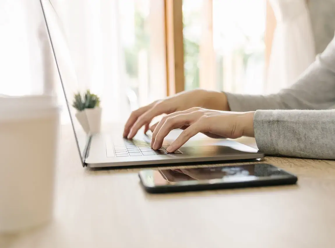 Close-up of hands typing on a laptop with a smartphone on the desk, representing online surety bond applications and digital insurance tools