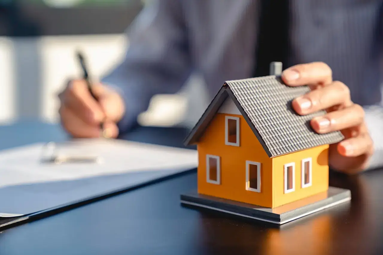 Close-up of a real estate agent signing mortgage documents with a miniature orange house model in the foreground, representing mortgage broker bonds and home loan agreements.
