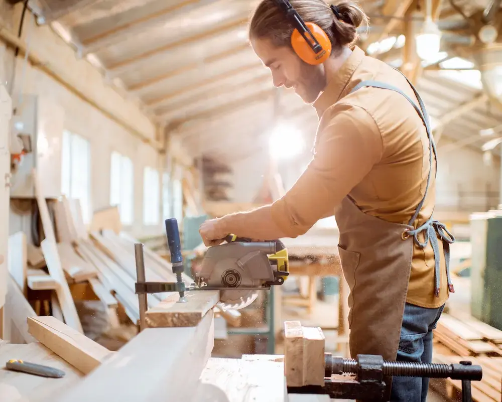 Male carpenter wearing ear protection while using a table saw in a woodshop.