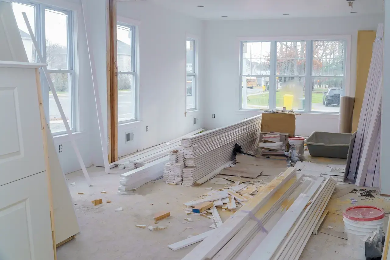Home interior under construction with stacked wood trim, drywall materials, and exposed windows during residential renovation