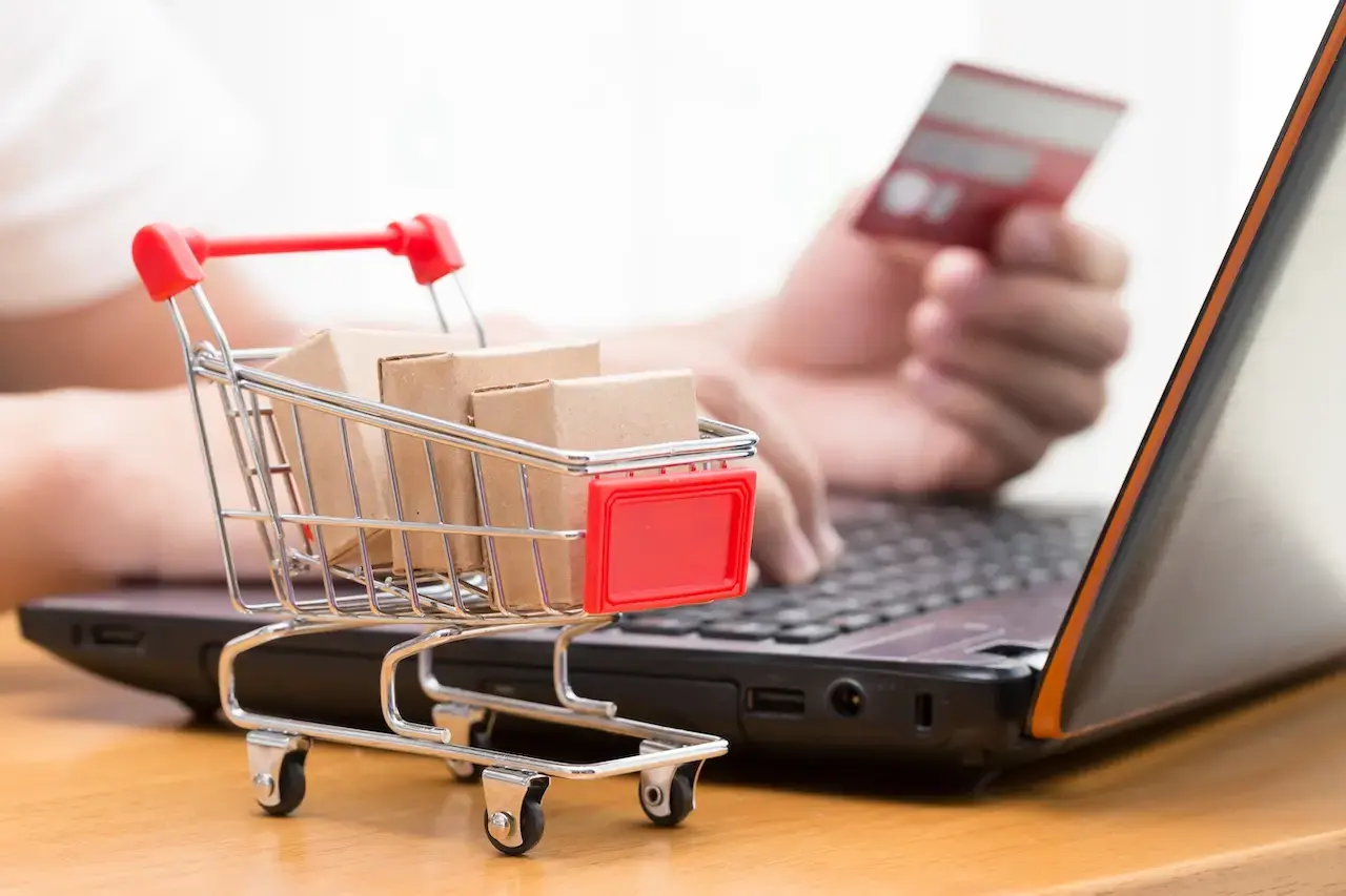 Close-up of a miniature shopping cart filled with cardboard boxes placed on a desk next to a laptop, representing e-commerce and online business setup.