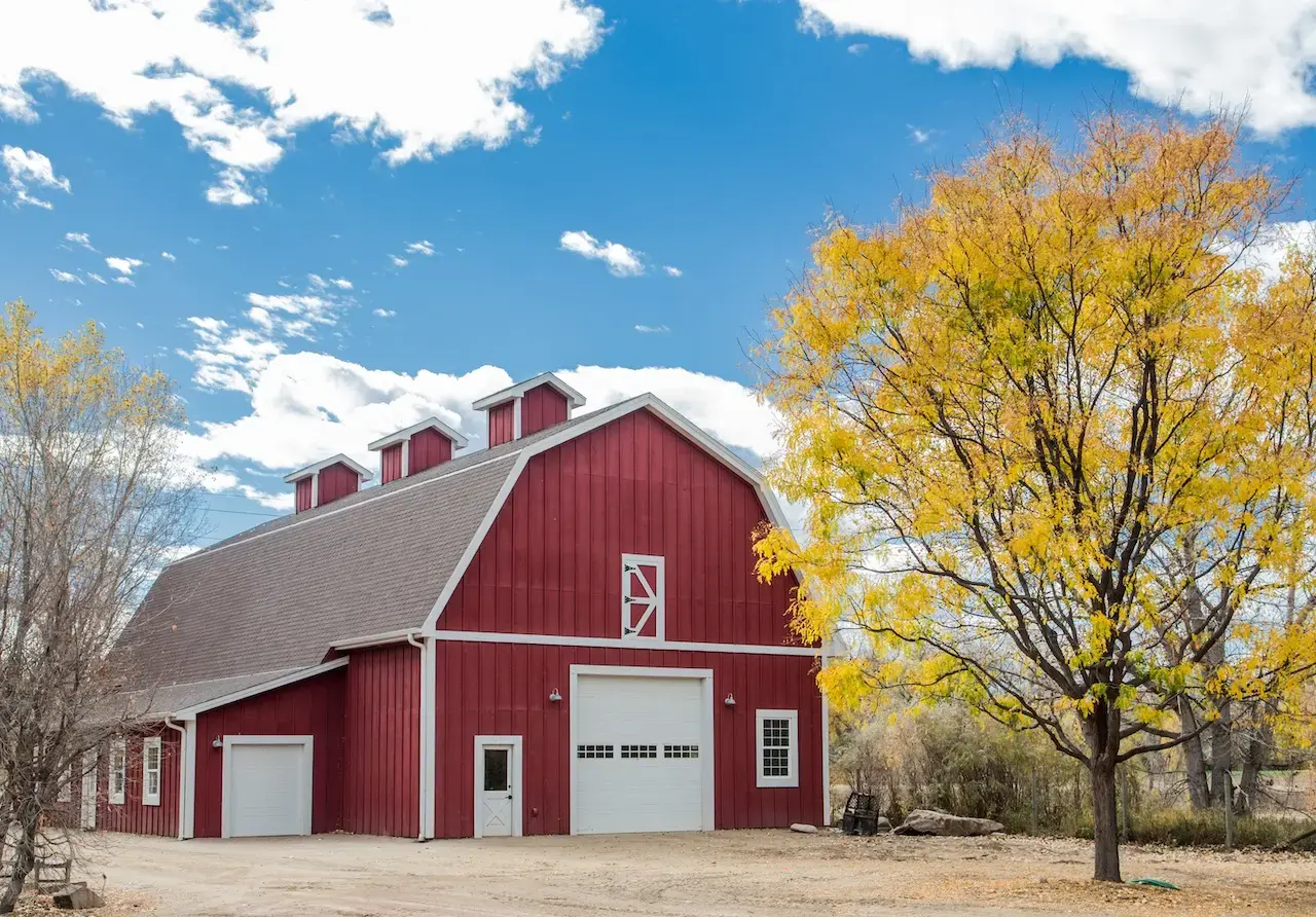 Large red barn with white trim and roll-up doors surrounded by autumn trees under a bright blue sky on a rural farm property.